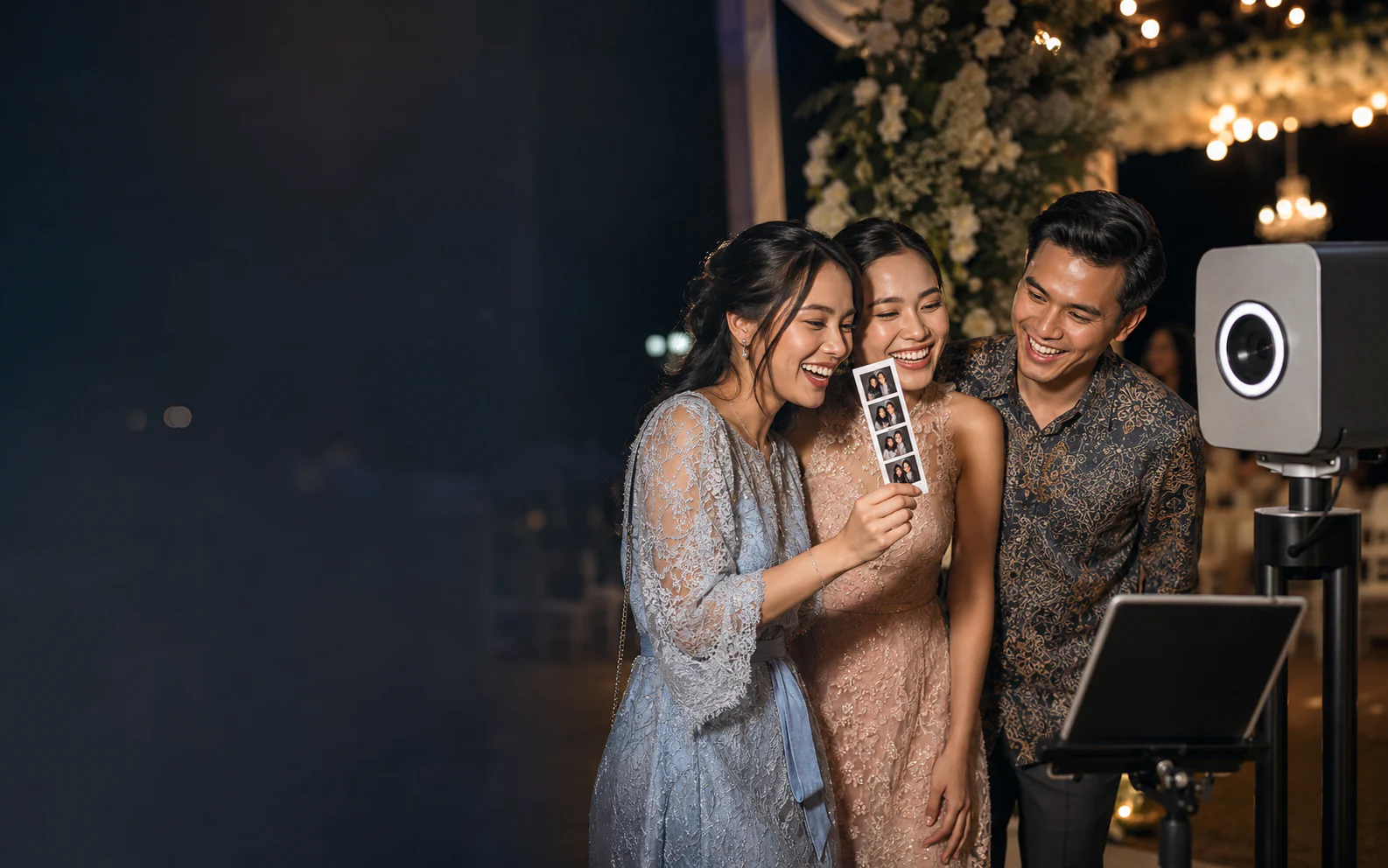 Tiga tamu tertawa sambil melihat hasil cetak photobooth di sebuah resepsi malam.